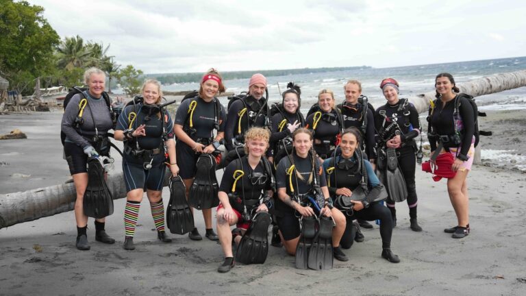 Scuba divers in wetsuits and gear pose on a sandy beach with palm trees and ocean behind them